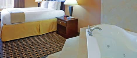 A hotel room with a large bed, a nightstand with a lamp, a bathroom with a bathtub, and a patterned rug.