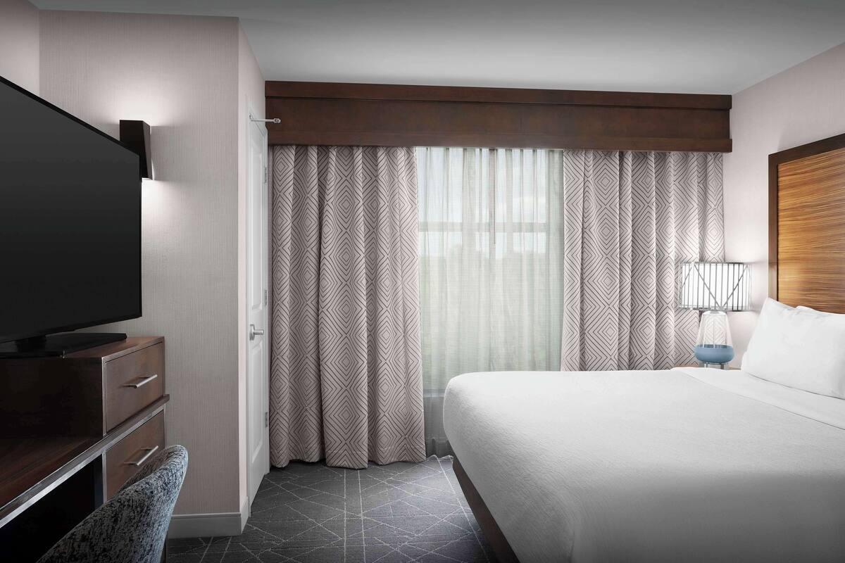 Ein Hotelzimmer mit einem großen Bett, einem Flachbildfernseher, einem Schreibtisch mit Schubladen und gemusterten Vorhängen.