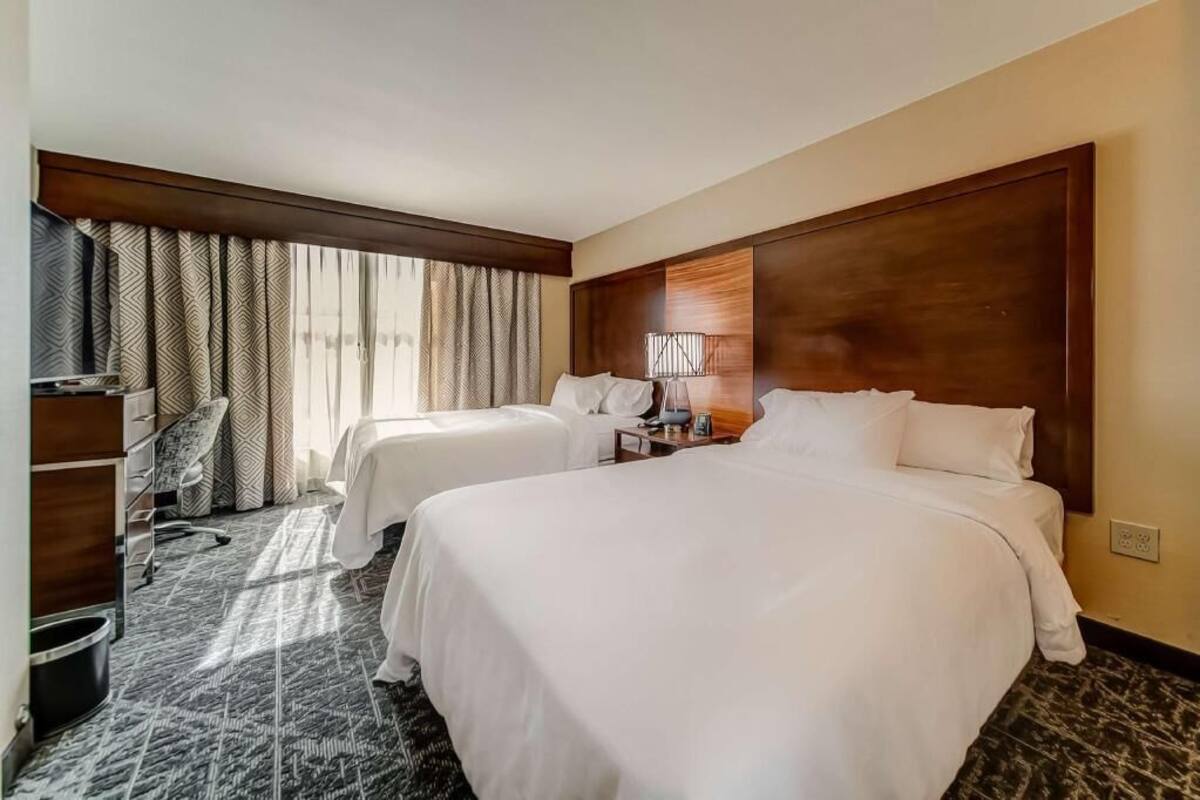 Ein Hotelzimmer mit zwei Betten, einem Schreibtisch, einem Stuhl und einem großen Fenster mit Vorhängen.