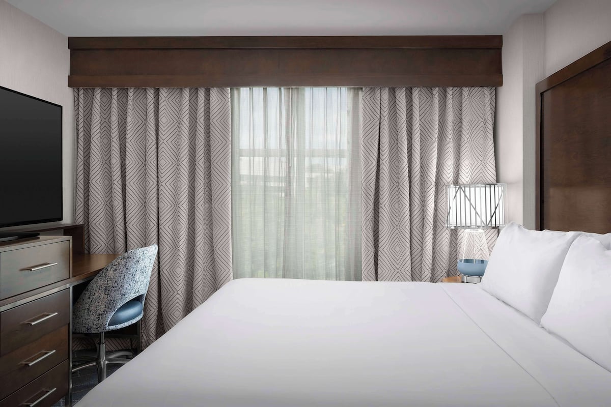 Ein Hotelzimmer mit einem großen Bett, einem Schreibtisch mit Stuhl, einem Fernseher und einem Fenster mit gemusterten Vorhängen.