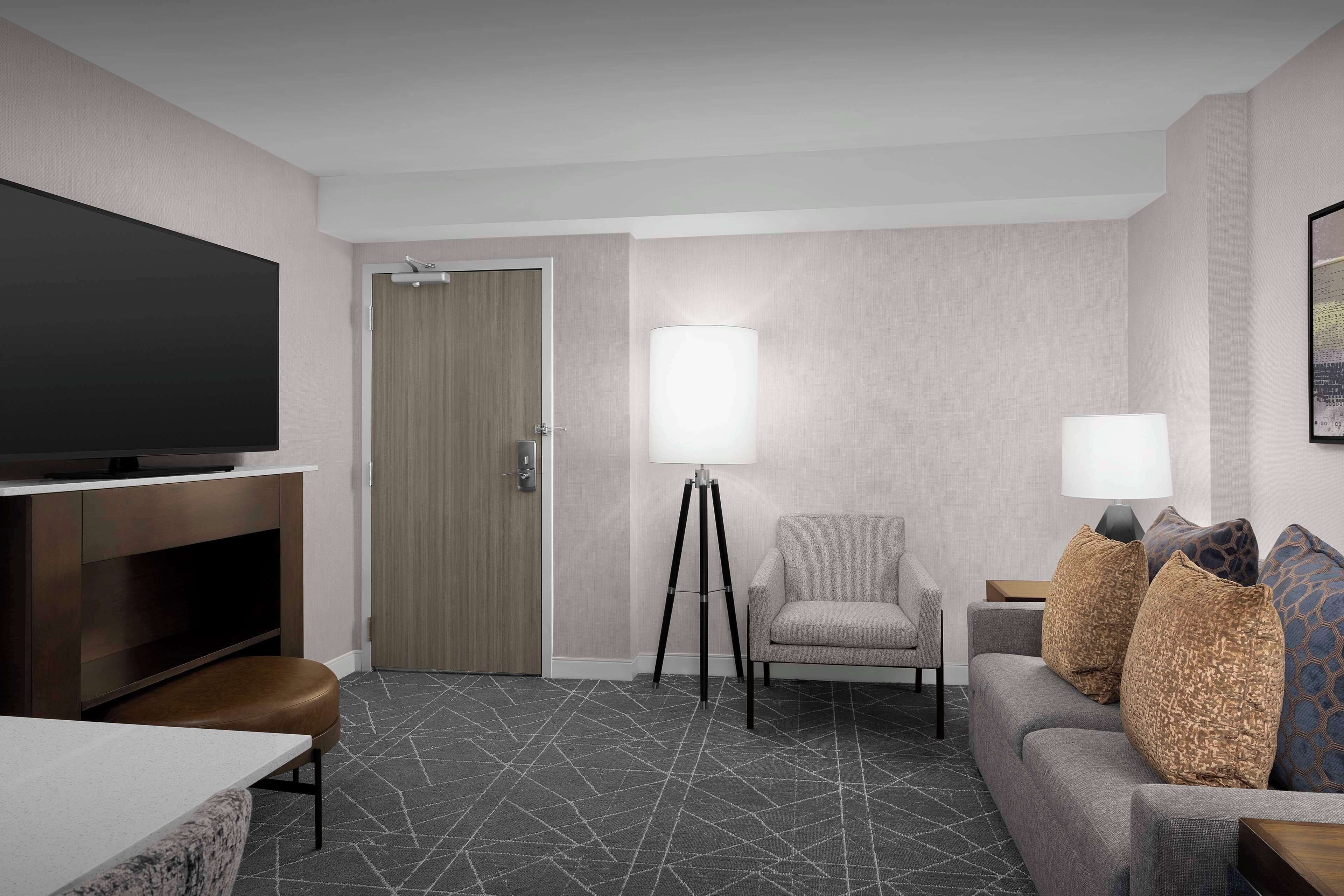 A hotel room with a flat-screen TV, a sofa, a chair, a lamp, and a patterned carpet.