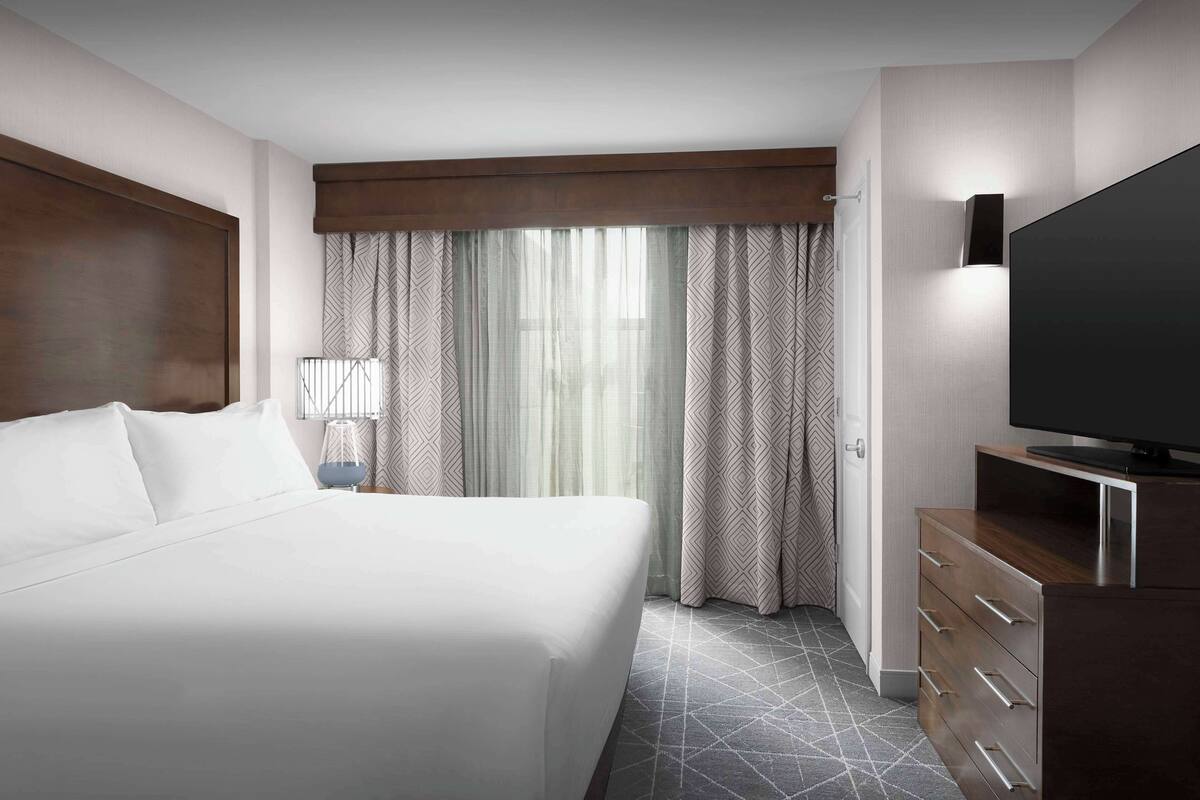 Ein Hotelzimmer mit einem großen Bett, einem Fernseher, einem Schminktisch und einem Fenster mit Vorhängen.