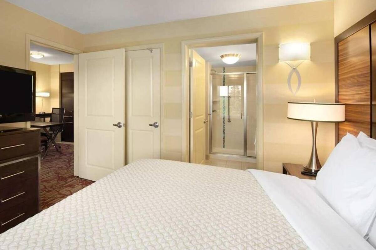 Ein Hotelzimmer mit einem Bett, einem Schreibtisch, einem Sessel, einem Fernseher, einer Lampe und einer Dusche.