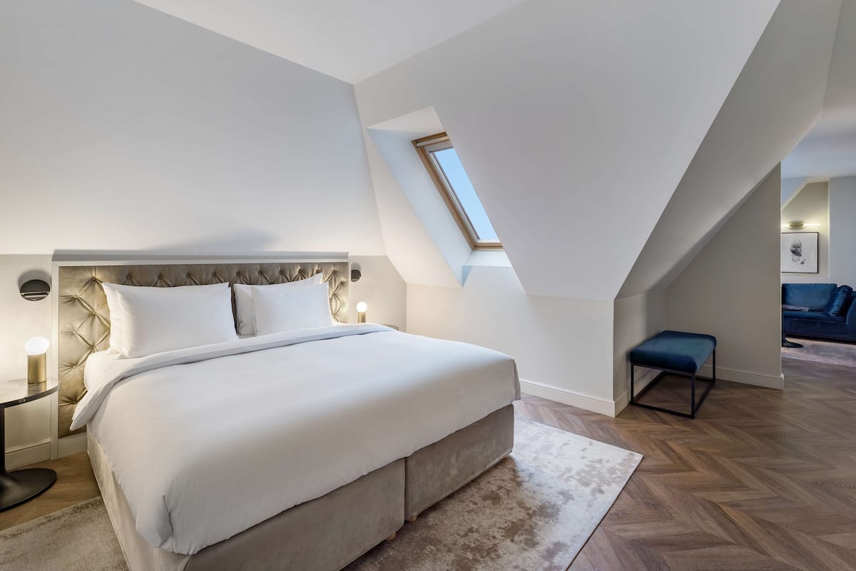 A modern bedroom with a large bed, a skylight, a bench, and a small table with a lamp.