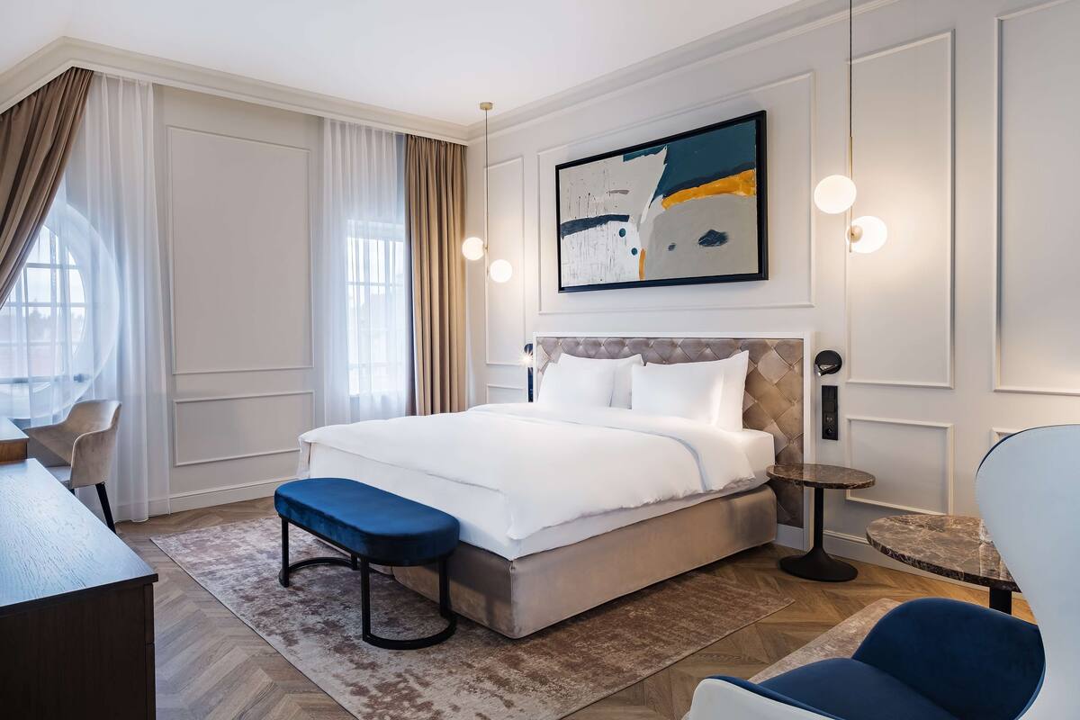 A modern hotel room with a large bed, a desk, a chair, and a small table.