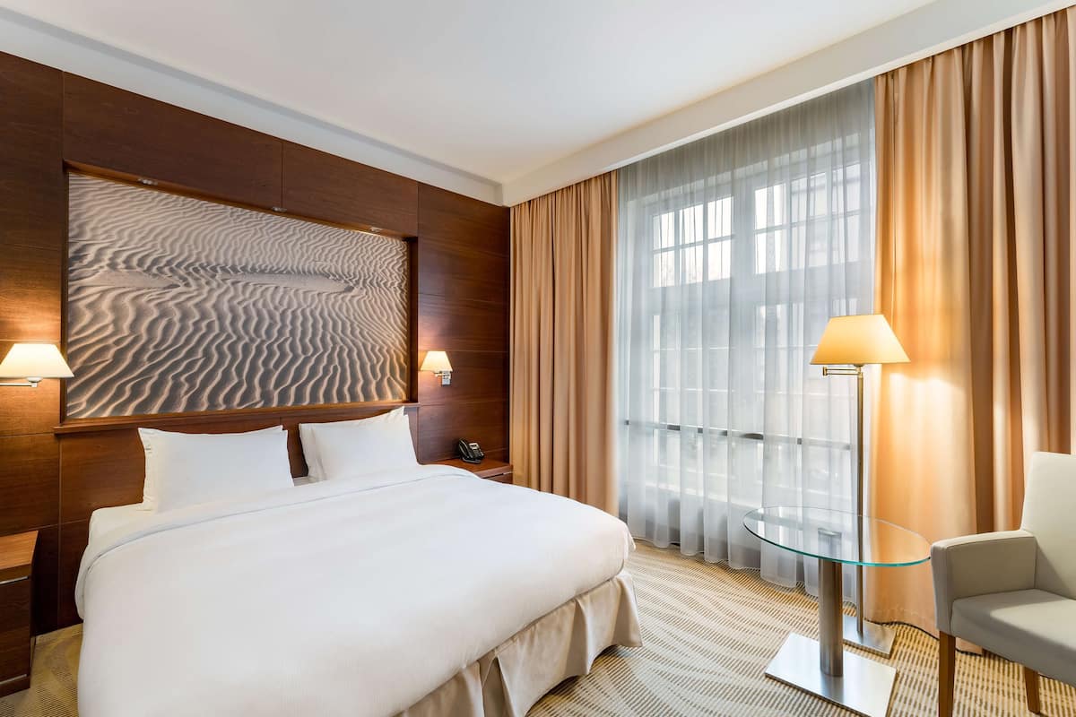 A hotel room with a large bed, a glass table, a chair, a wall with a textured design, and a window with curtains.