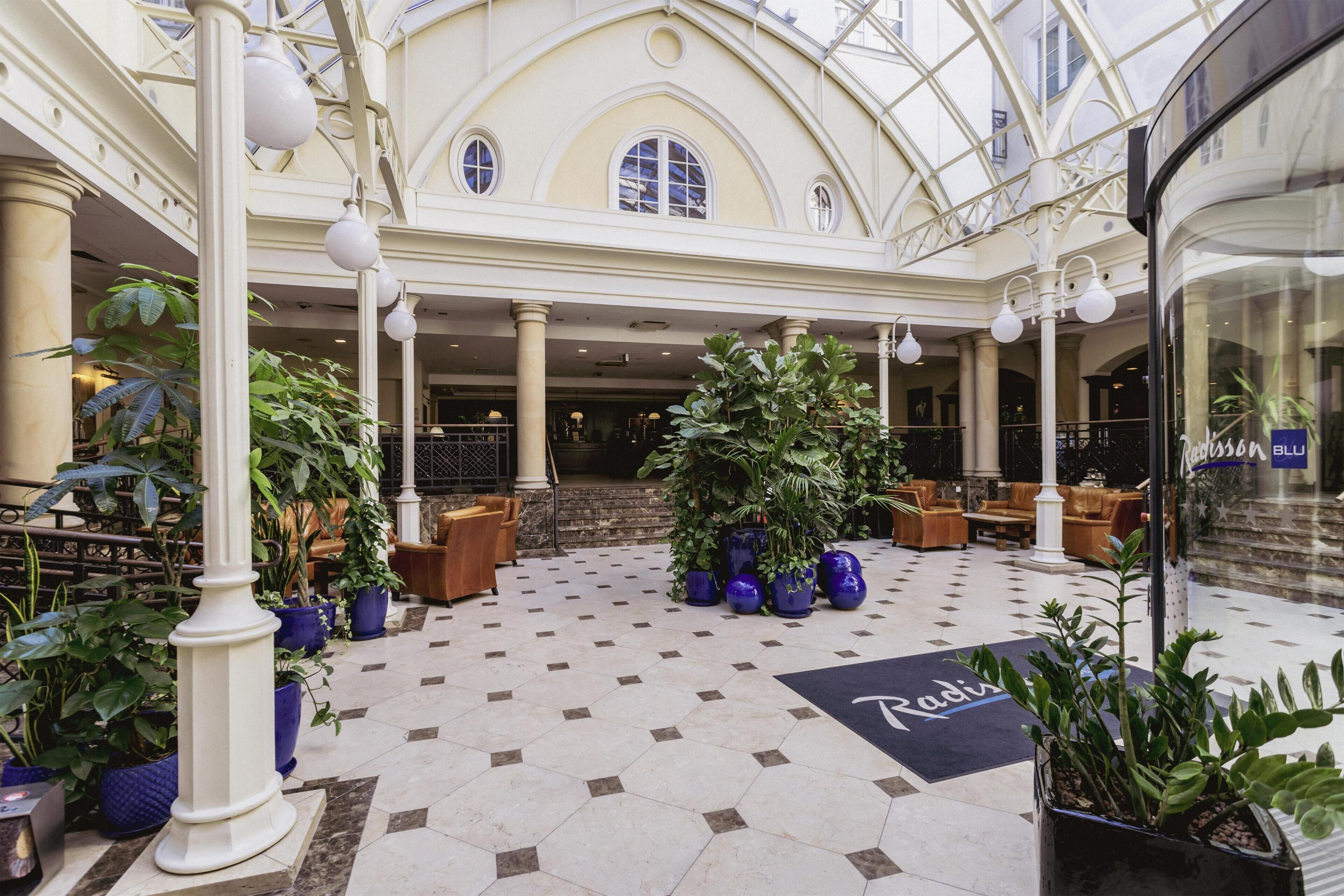 A spacious lobby with a high ceiling, large windows, and a central seating area with sofas and potted plants.