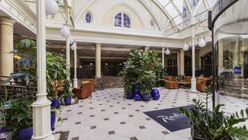 A spacious lobby with a high ceiling, large windows, and a central seating area with sofas and potted plants.