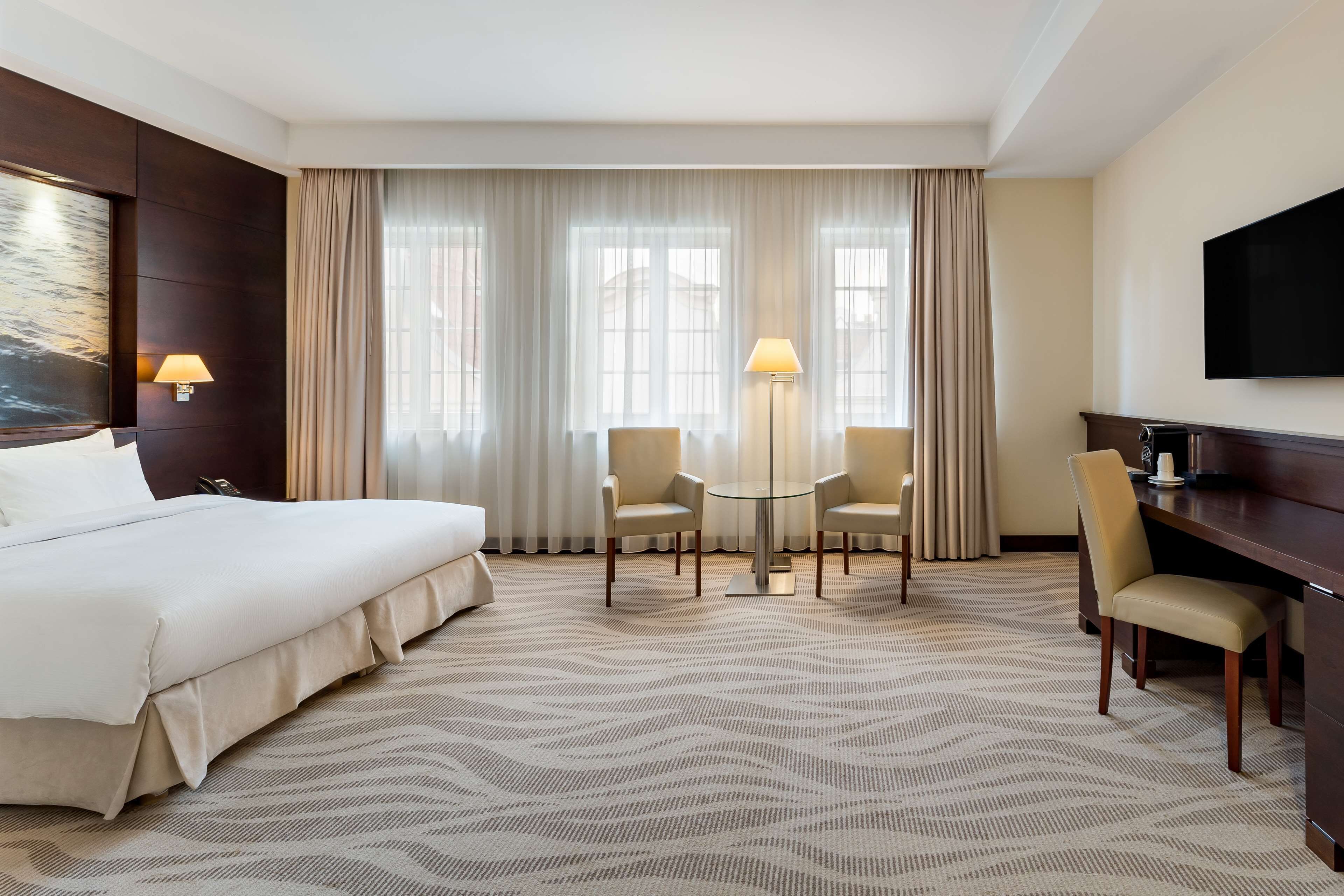 A hotel room with a large bed, a desk with a chair, a small table with two chairs, and a TV on the wall.