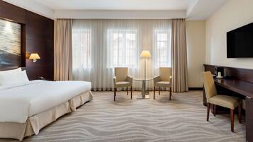 A hotel room with a large bed, a desk with a chair, a small table with two chairs, and a TV on the wall.