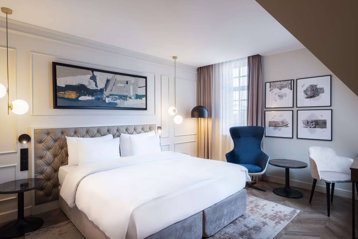 A modern hotel room with a large bed, a tufted headboard, a round table, a blue armchair, and a small round table.