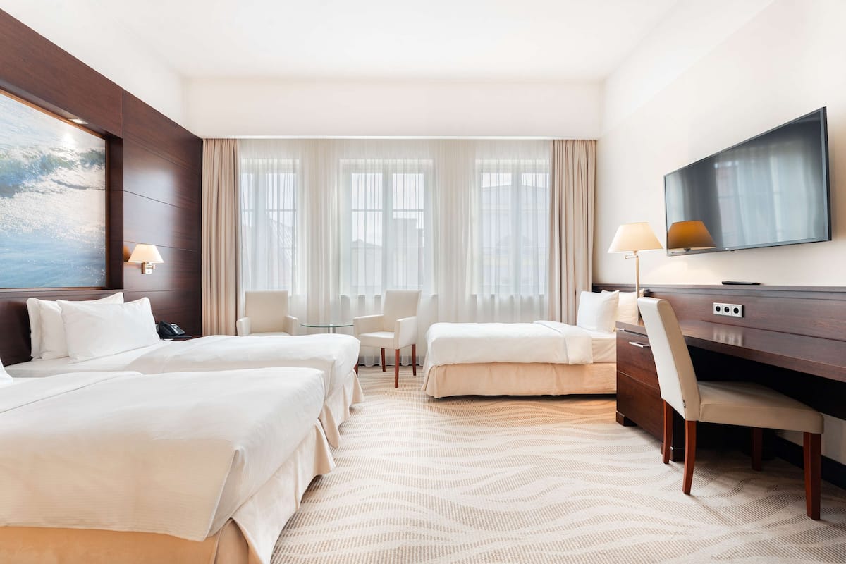 A hotel room with two beds, a desk, a chair, a TV, and a large window with curtains.