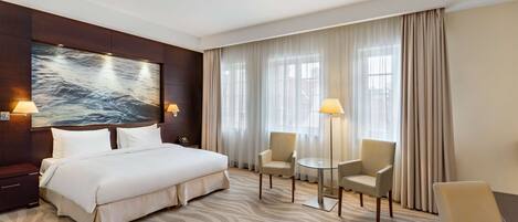 A hotel room with a large bed, two armchairs, a small round table, a wall-mounted TV, and a large window with curtains.