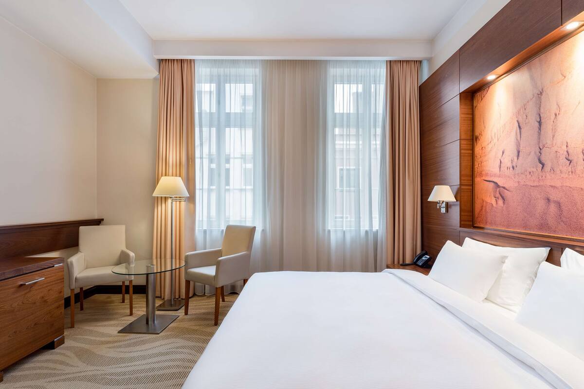 A hotel room with a large bed, two armchairs, a small table, a lamp, and a wooden headboard.