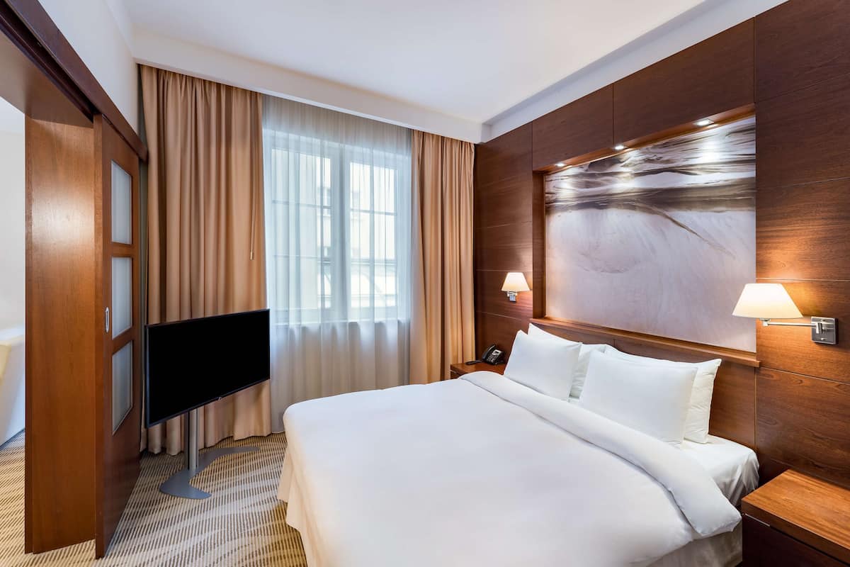 A hotel room with a large bed, a flat-screen TV, a wooden headboard, and a large window with curtains.