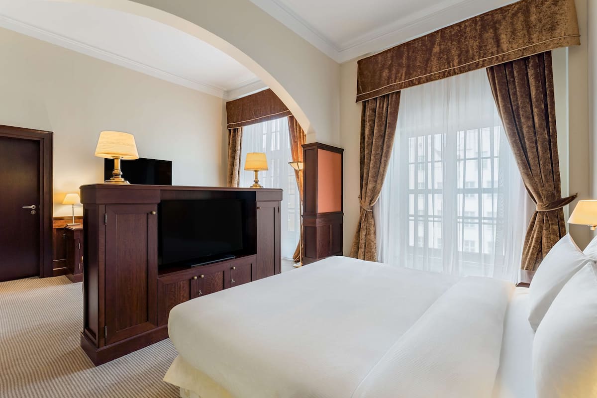 A hotel room with a large bed, a flat-screen TV, wooden furniture, and large windows with curtains.