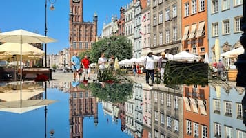 A historic European town square with a prominent clock tower, reflecting buildings, and a calm water body.