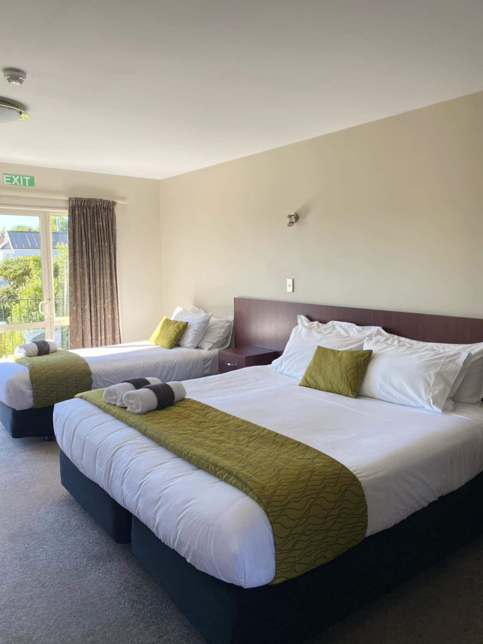 Ein Hotelzimmer mit zwei Betten, einem fensterbelagten Fenster, einem Fluchtwegweiser und Blick auf Grün.
