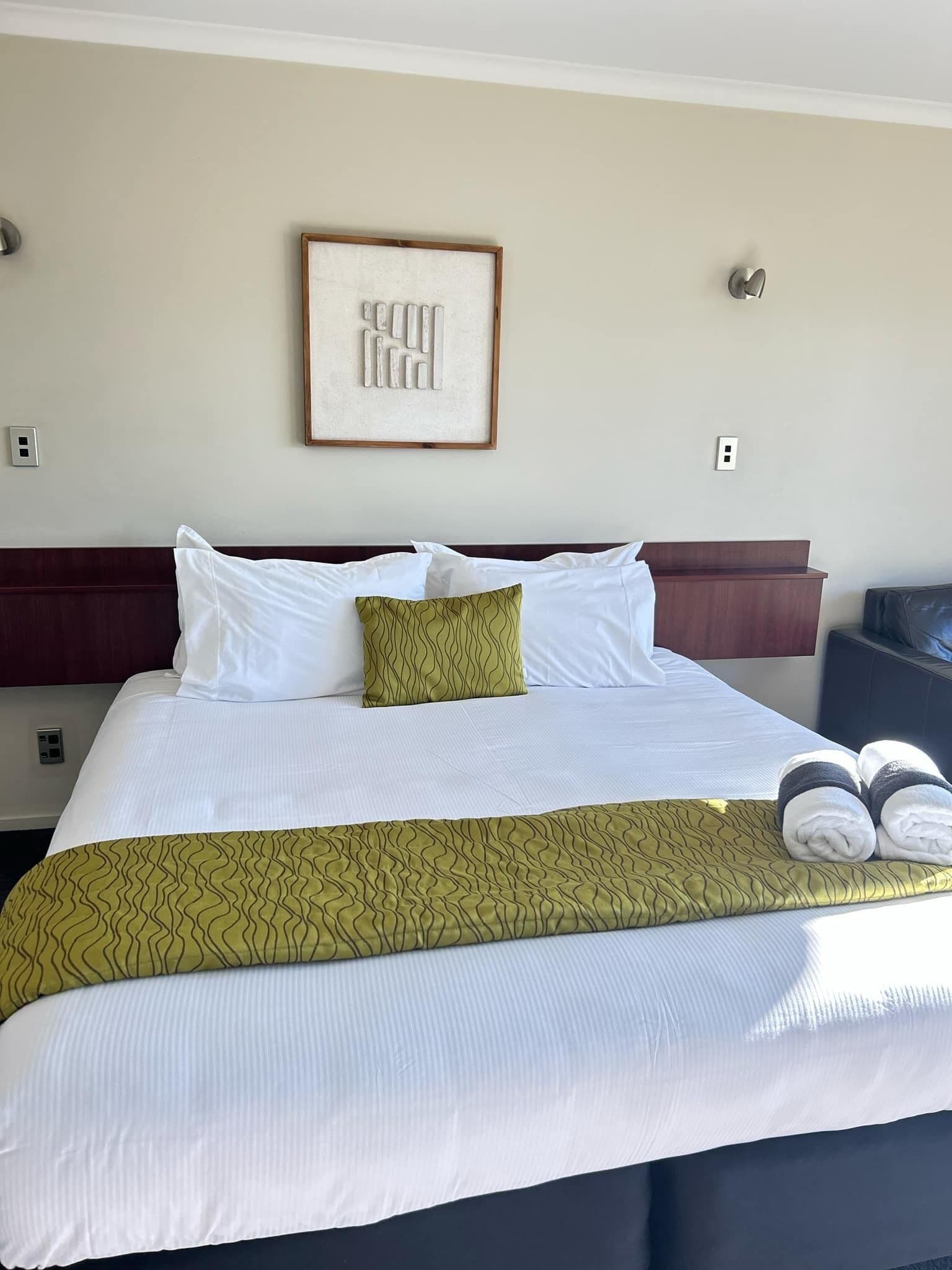 A neatly made bed with white and green bedding, a framed artwork above it, and a dark-colored nightstand to the right.