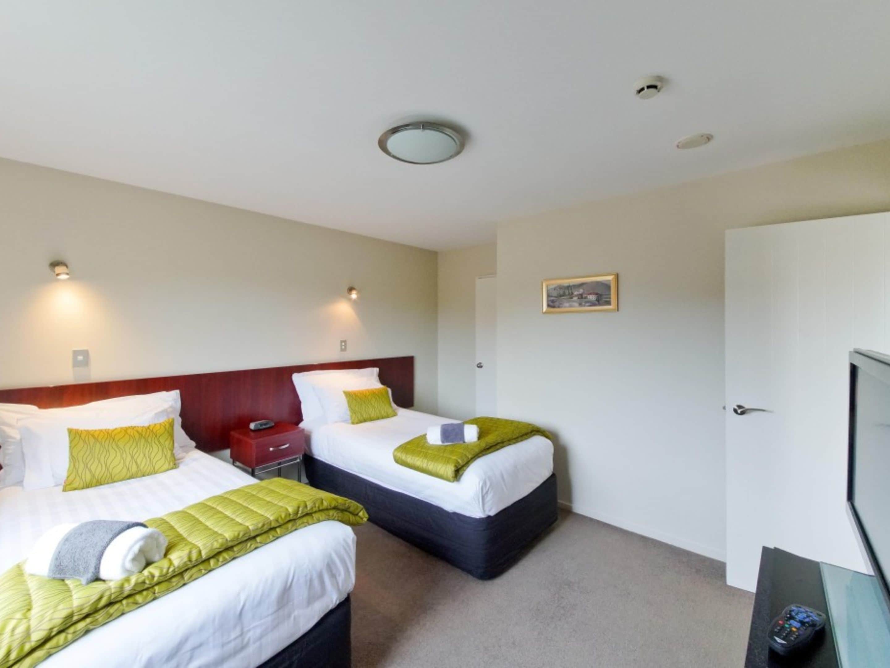 Une chambre d’hôtel avec deux lits, chacun agrémenté de draps verts et blancs, une table de chevet rouge, un téléviseur et une porte.