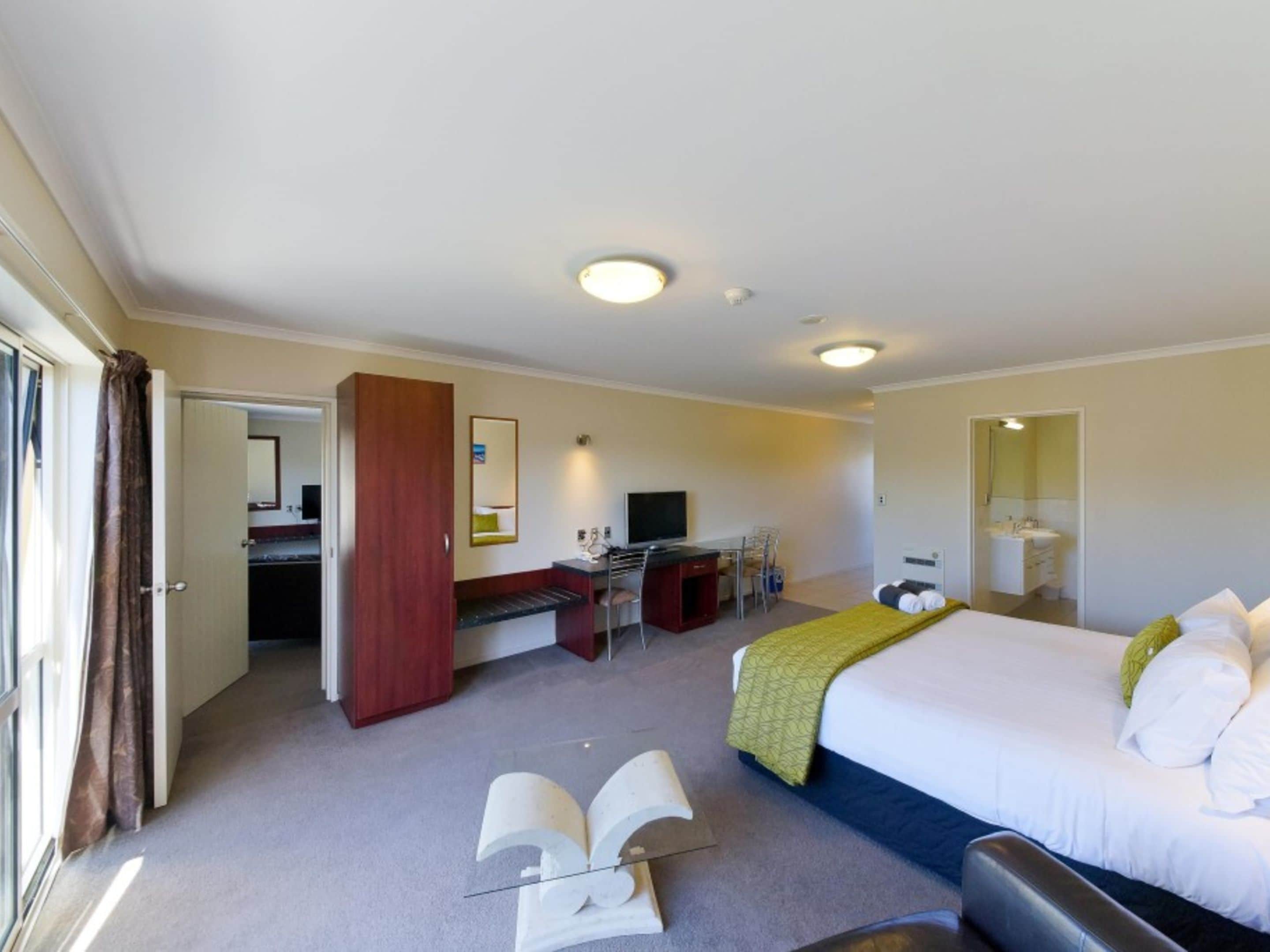 A hotel room with a bed, a desk, a chair, a television, and a bathroom visible through an open door.