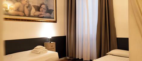 Quarto de hotel com duas camas, um quadro na parede, uma mesa de cabeceira com abajur e cortinas na janela.