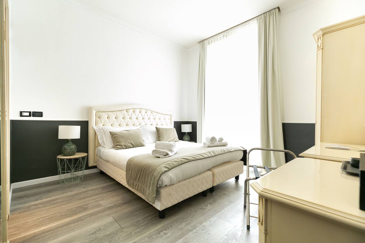 A hotel room with a bed, bedside tables, a desk, and a large mirror.