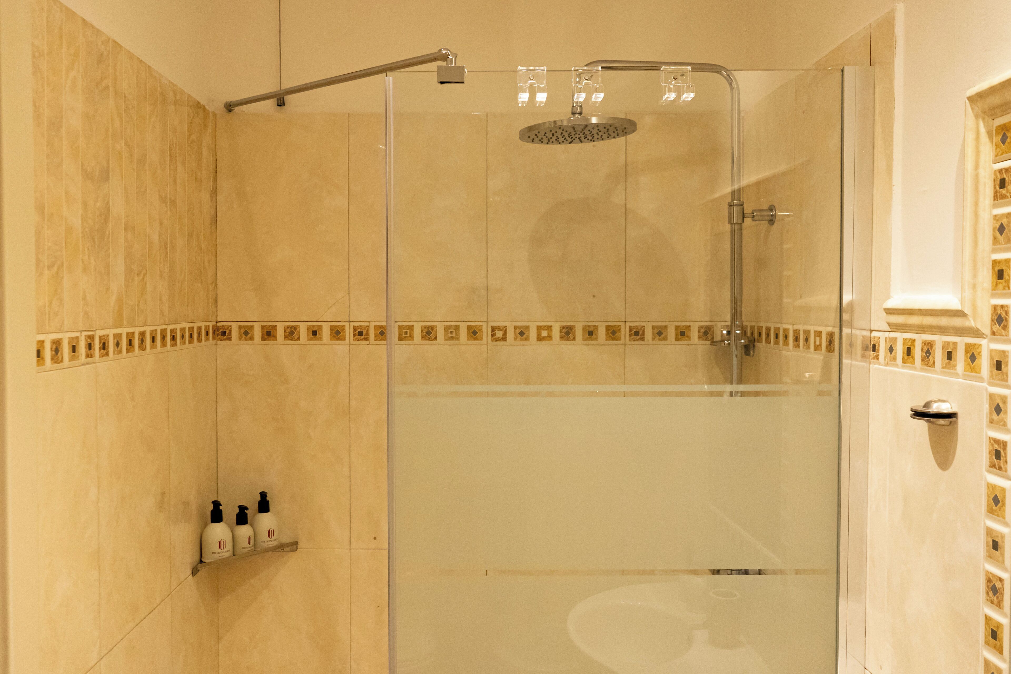 Una ducha envuelta en vidrio con cabezal de ducha de mano y otro cabezal de ducha fijo en la pared. Hay tres botellas en un estante a la izquierda de la ducha.