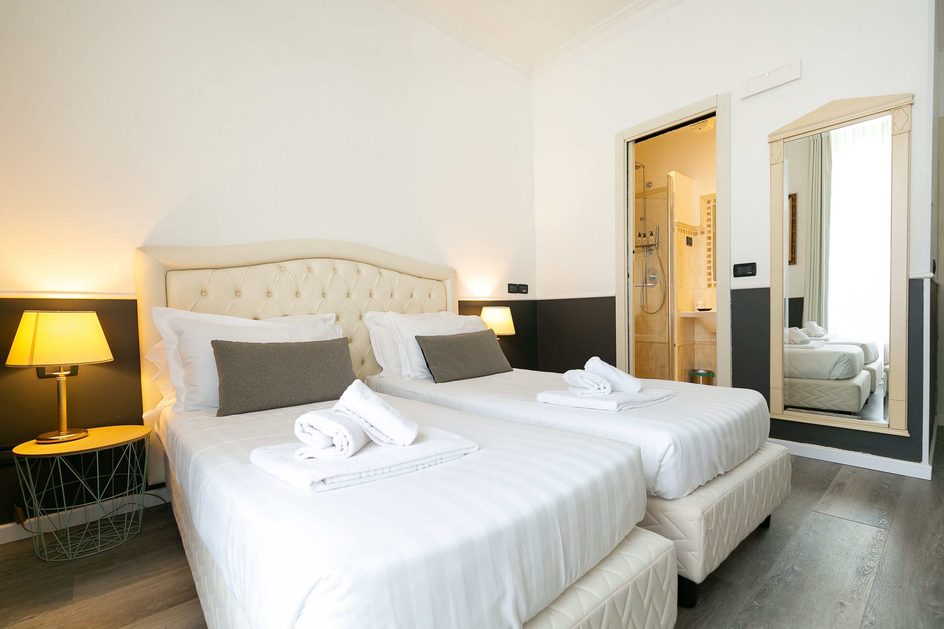 A hotel room with two beds, a nightstand with a lamp, a bathroom visible through an open door, and a large mirror.