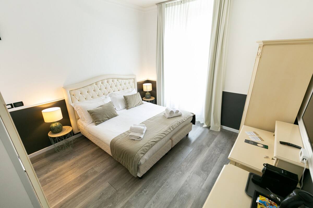 A hotel room with a large bed, bedside tables with lamps, a desk with a computer, and a window with curtains.