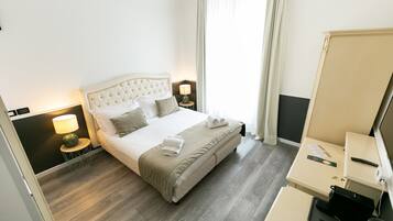 A hotel room with a large bed, bedside tables with lamps, a desk with a computer, and a window with curtains.