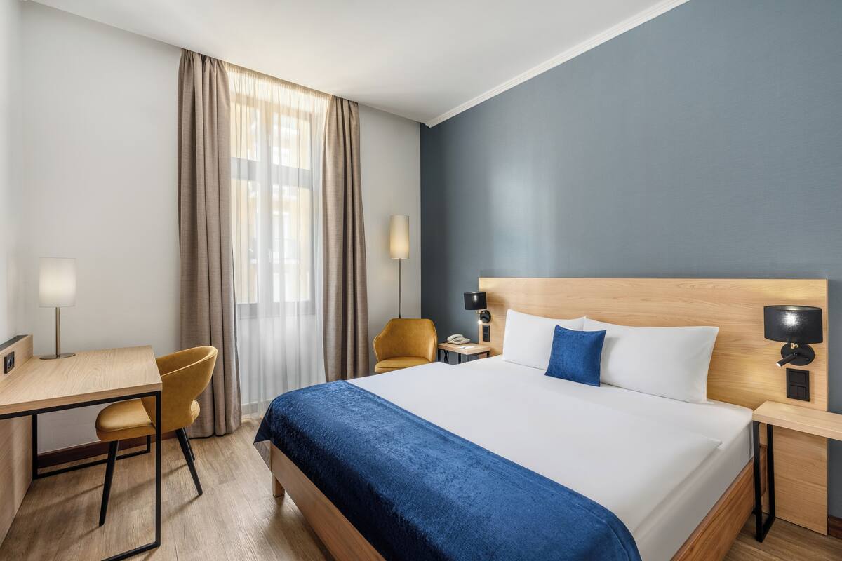 A hotel room with a bed, a desk, a chair, a lamp, and a window with curtains.