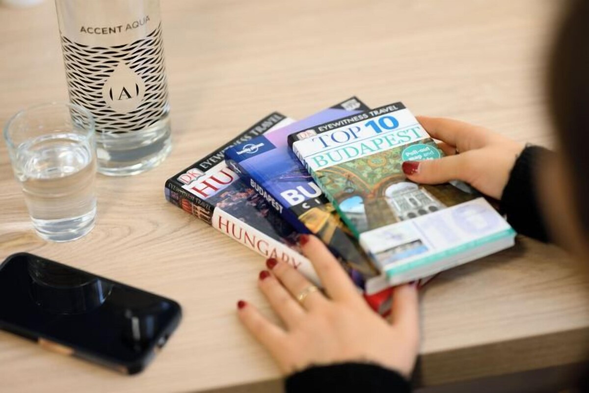 A person holding a book titled 'Top 10 Budapest' with other books on a table, a glass of water, and a bottle labeled 'Accent Aqua.'