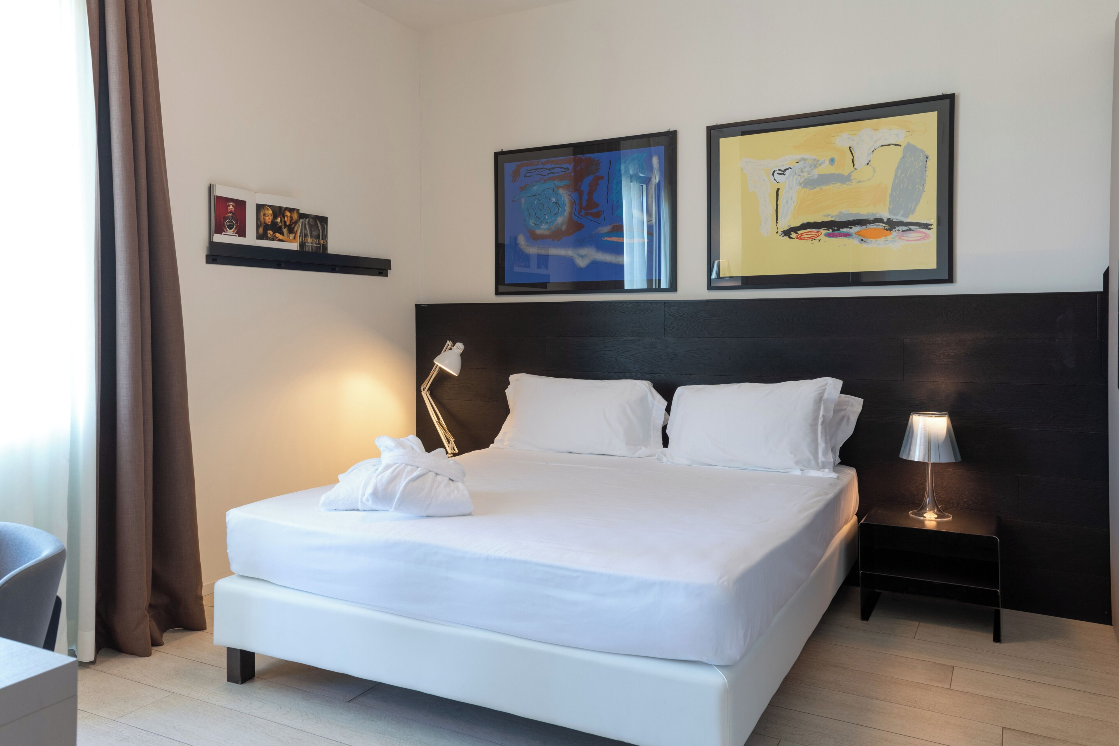 Una cama bien hecha con sábanas blancas, una lámpara de noche y dos cuadros abstractos en la pared.