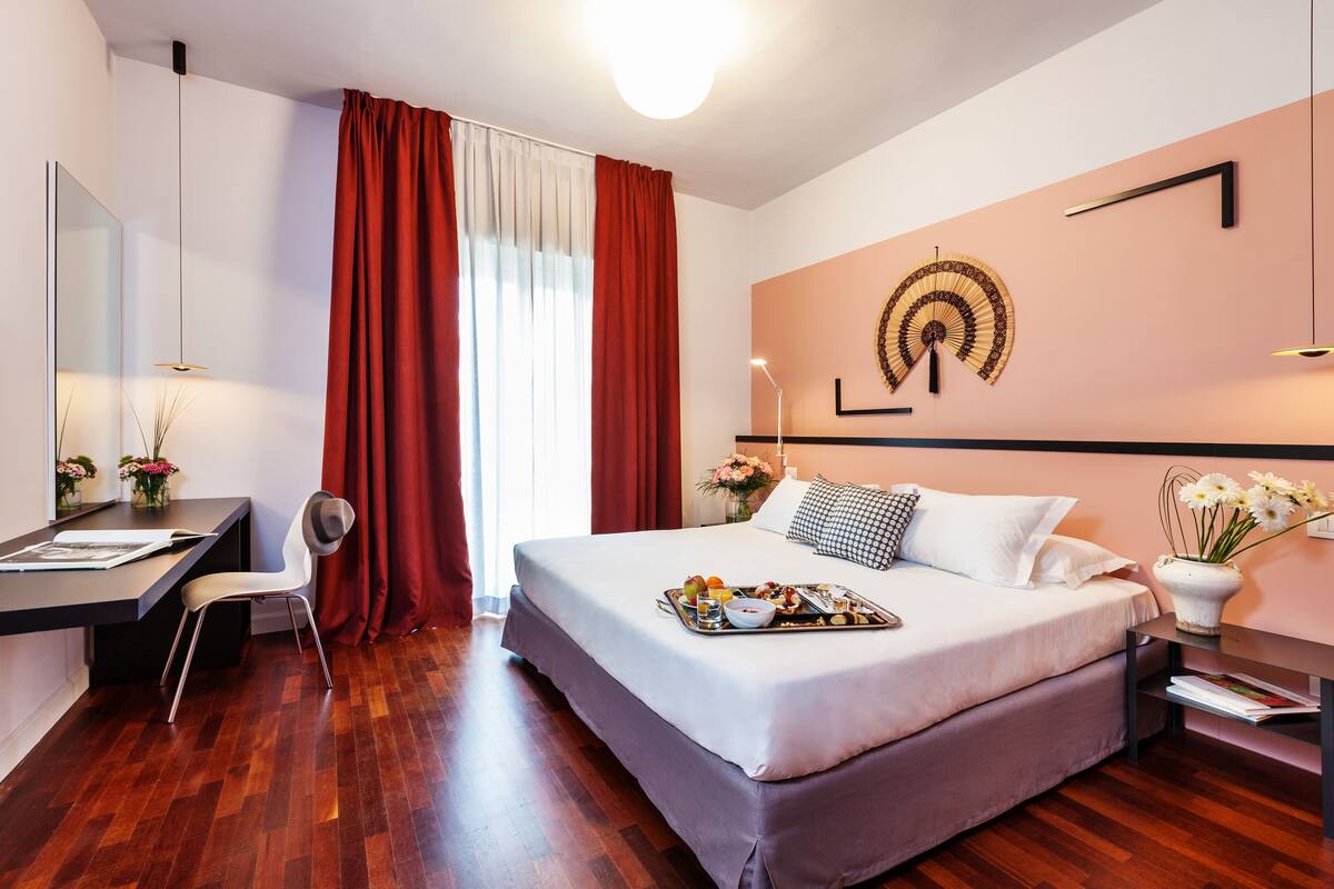 A modern hotel room with a large bed, a desk with a chair, a mirror, and a wall-mounted fan.