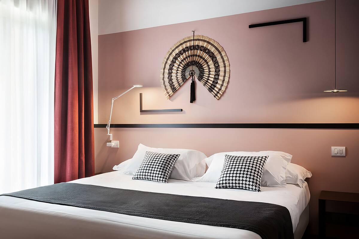 A neatly made bed with white linens and black and white patterned pillows, a wall-mounted decorative fan, and a bedside lamp.