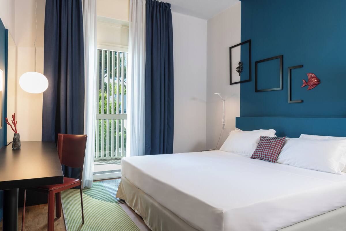 A hotel room with a bed, a desk, and a chair. There is a balcony with curtains and a view of greenery.