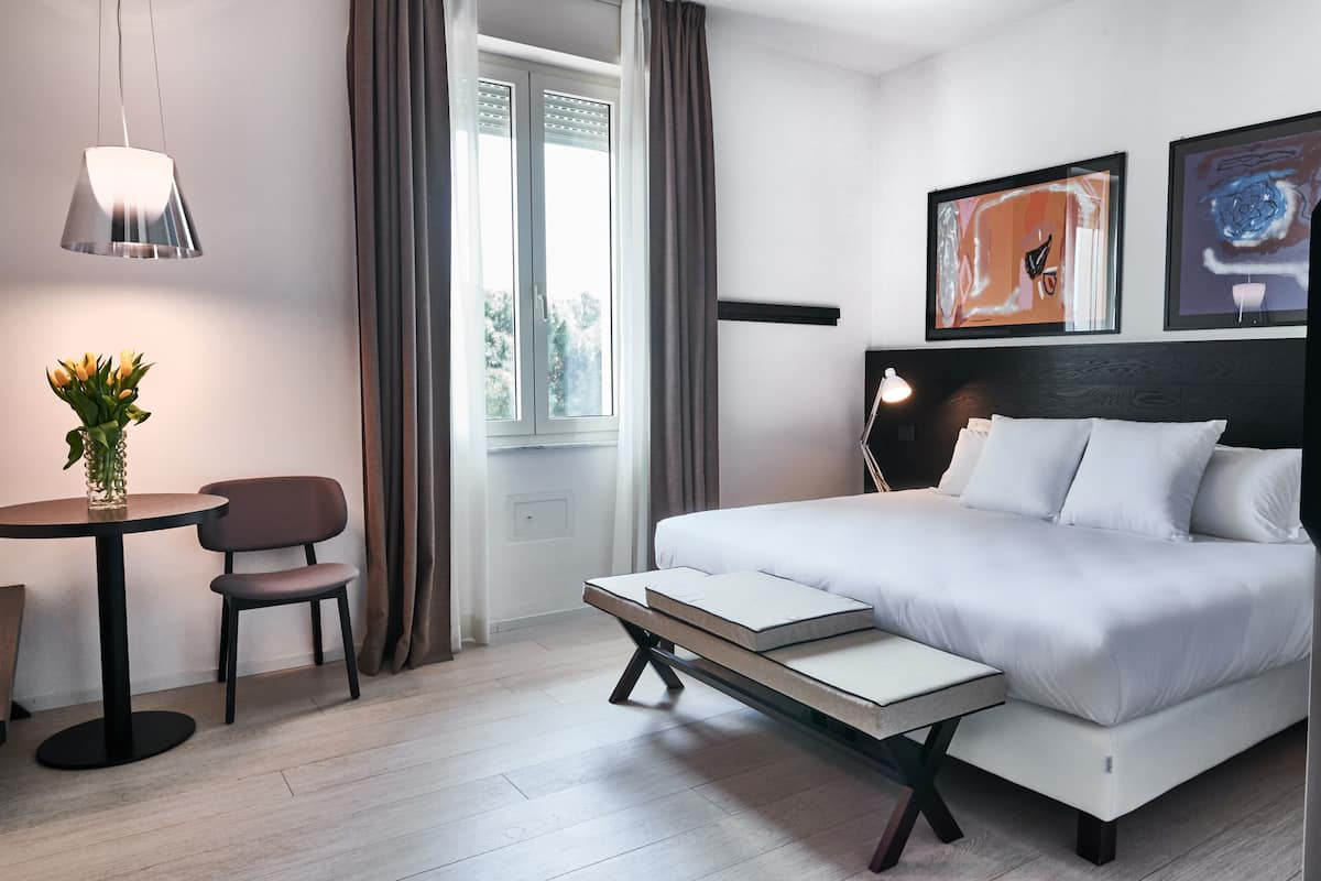 A modern hotel room with a bed, bedside table, chair, and a small table with a vase of flowers.