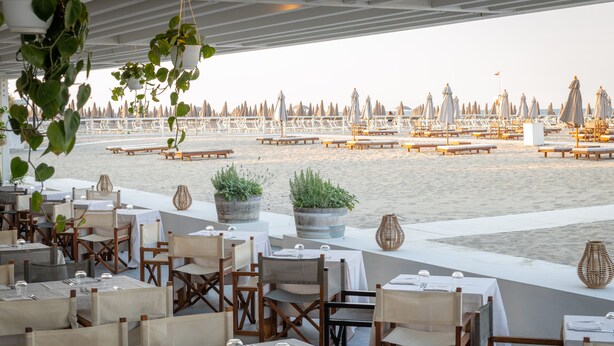 Een terras met tafels en stoelen, met uitzicht op een strand met parasols en ligbedden.