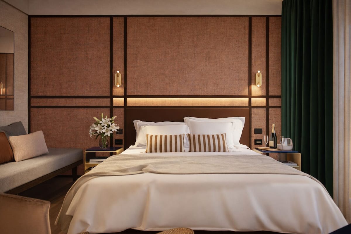 A hotel room with a large bed, a bedside table with a vase of flowers, a nightstand with a bottle and glasses, and a green curtain.