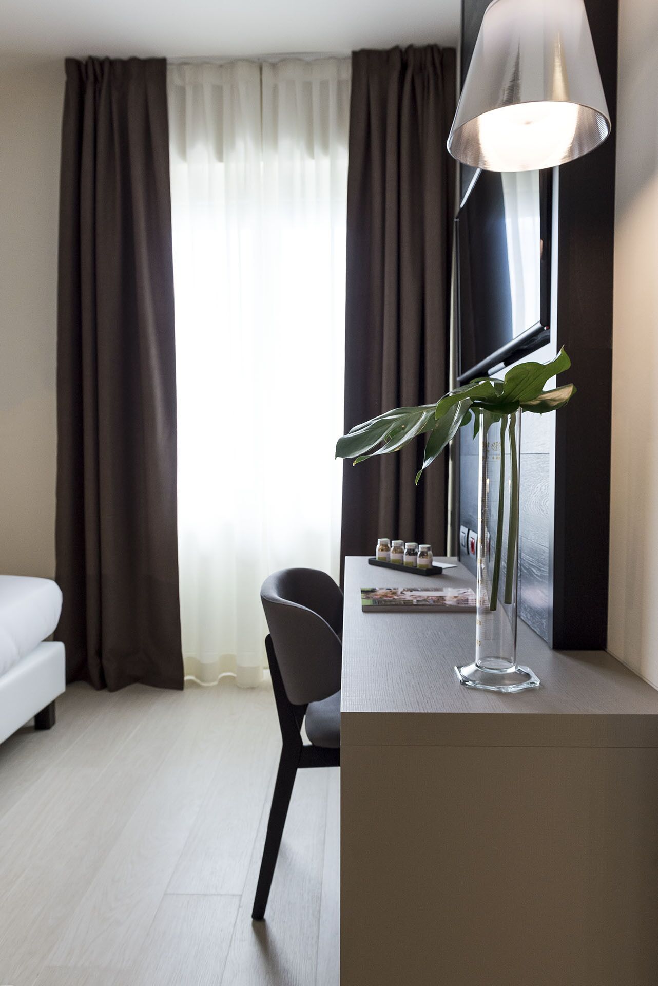 Een hotelkamer met een bed, een bureau met een lamp, een stoel, een vaas met een plant en een raam met gordijnen.