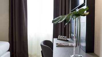 Een hotelkamer met een bed, een bureau met een lamp, een stoel, een vaas met een plant en een raam met gordijnen.