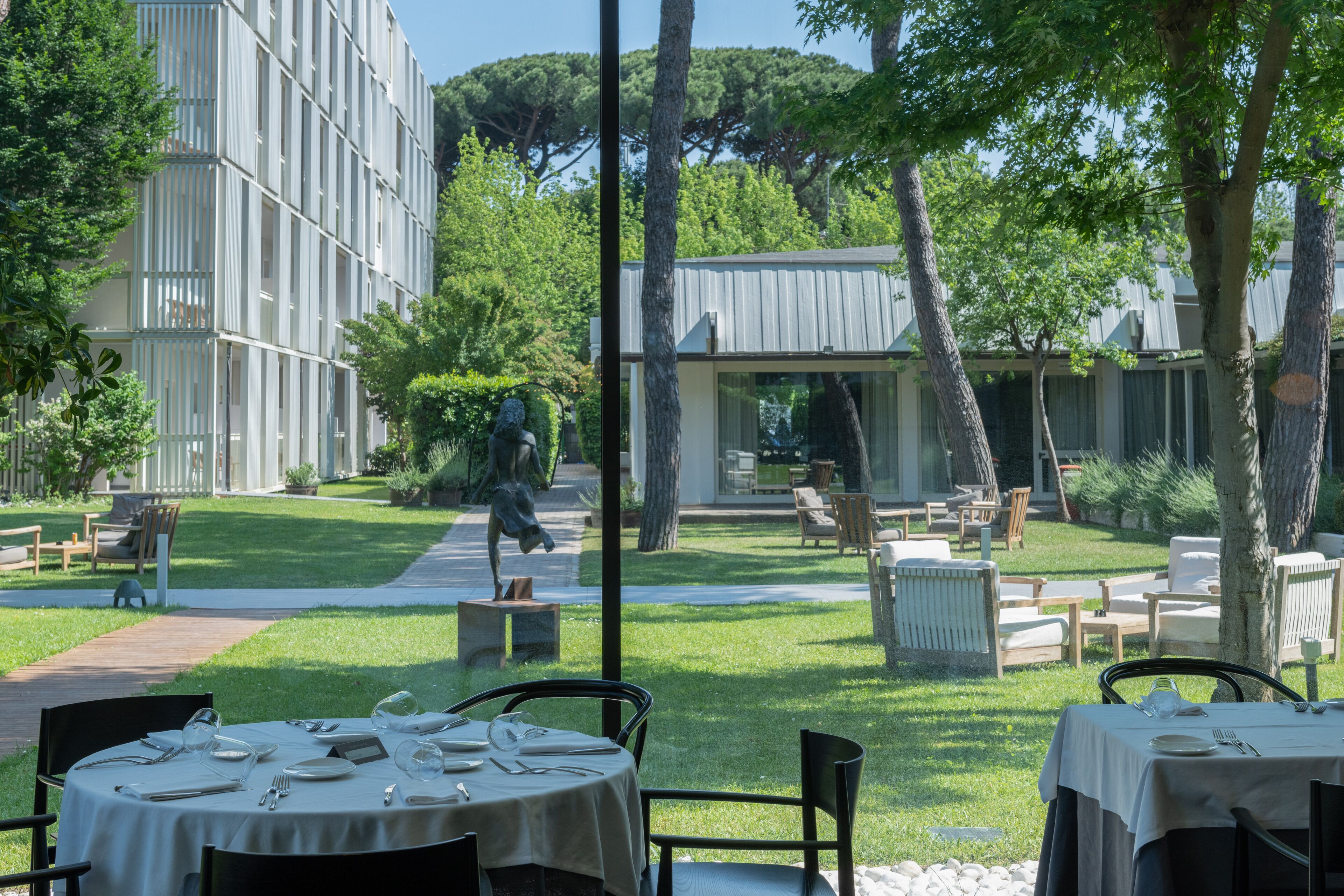 Un espace de restauration en plein air, avec des tables dressées pour un repas, entouré d’arbres et bordé d’un bâtiment au design moderne.