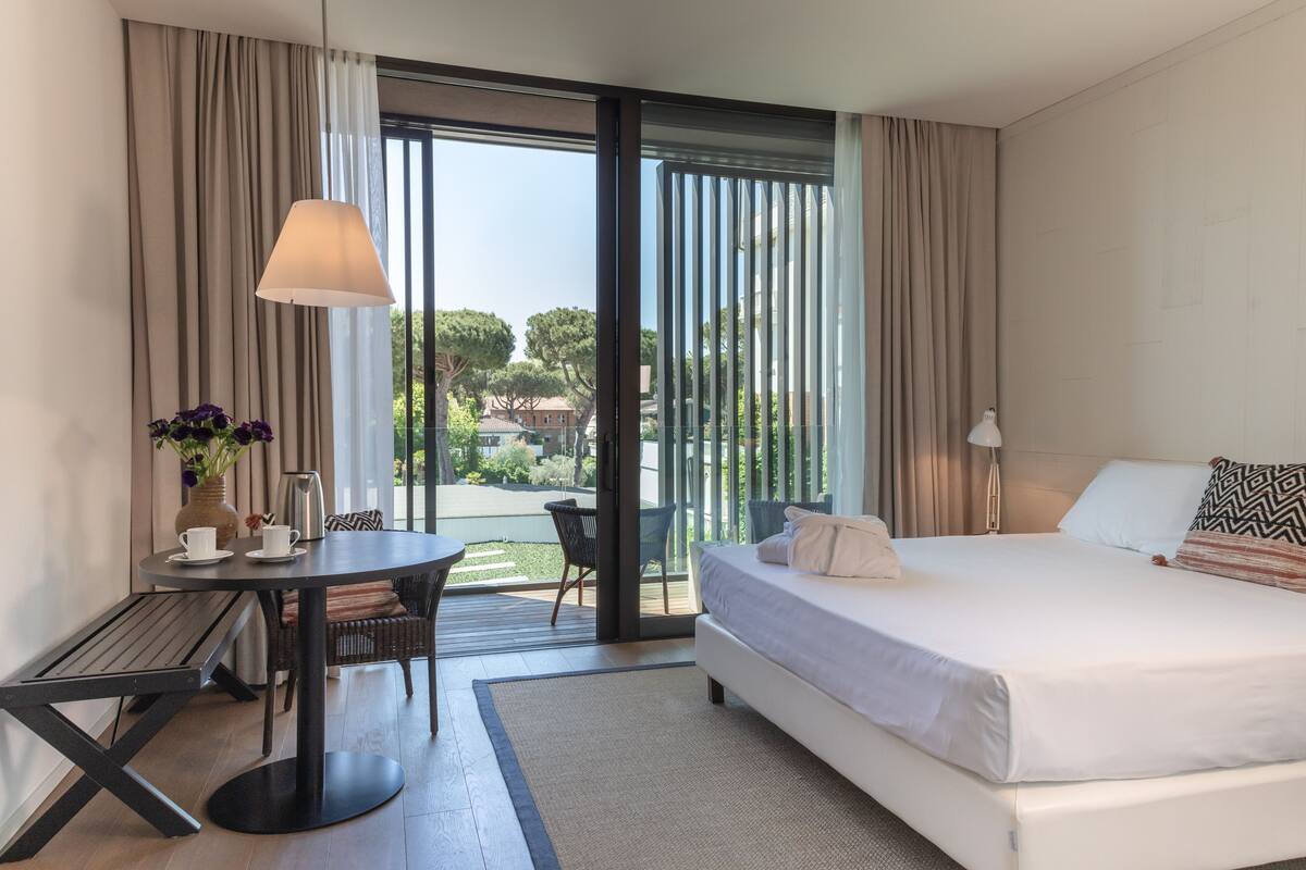 A modern hotel room with a large bed, a dining table, and a view of the outdoors.