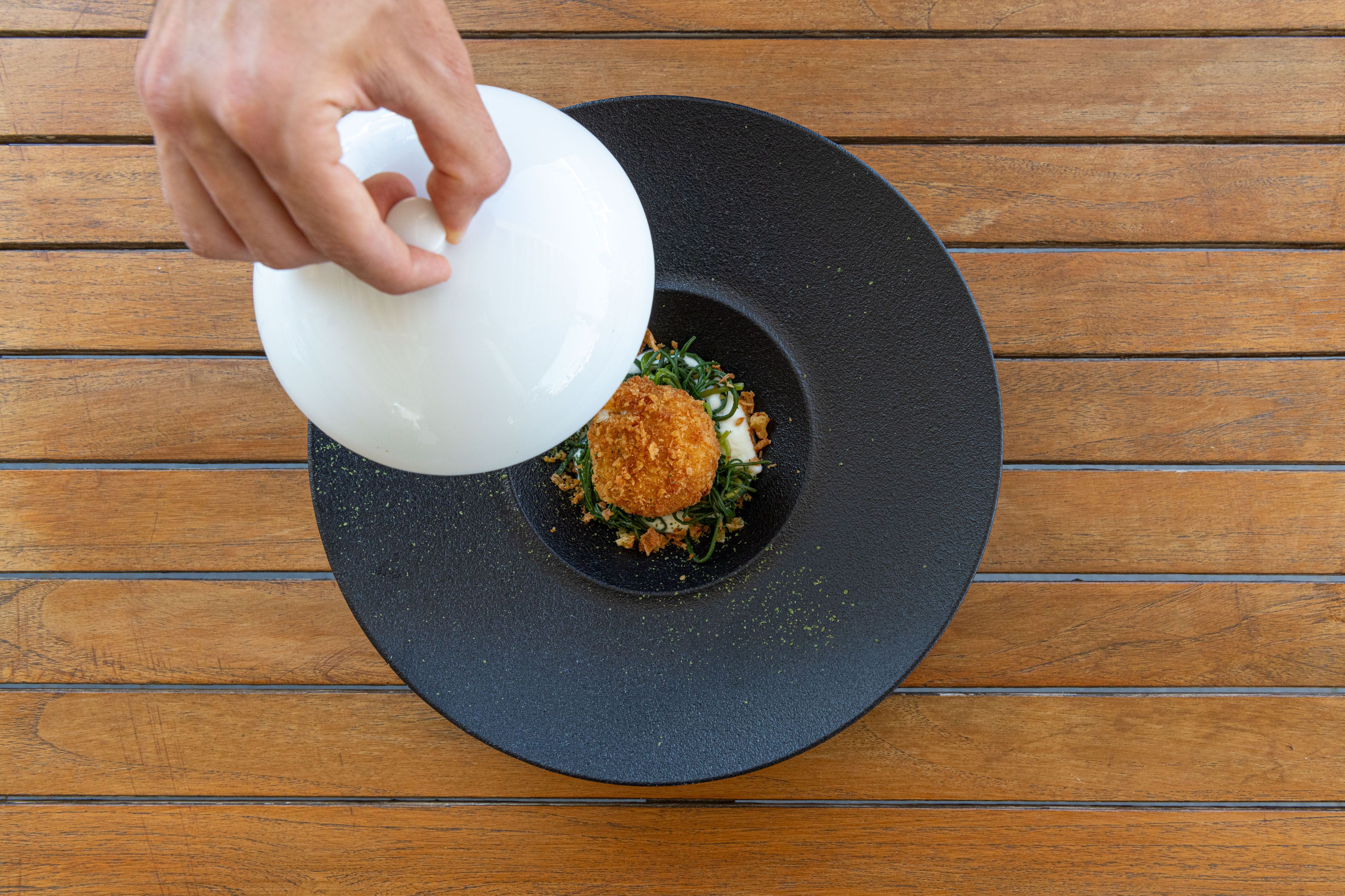 Una mano vertiendo un líquido blanco sobre un plato negro con comida.