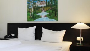 Ein Hotelzimmer mit Bett, weißen Kissen, einem Nachttisch mit Lampe und einem Gemälde einer Berglandschaft an der Wand.