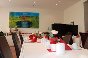Un comedor con mesas dispuestas para una comida, un cuadro grande en la pared y un televisor de pantalla plana sobre un mueble.