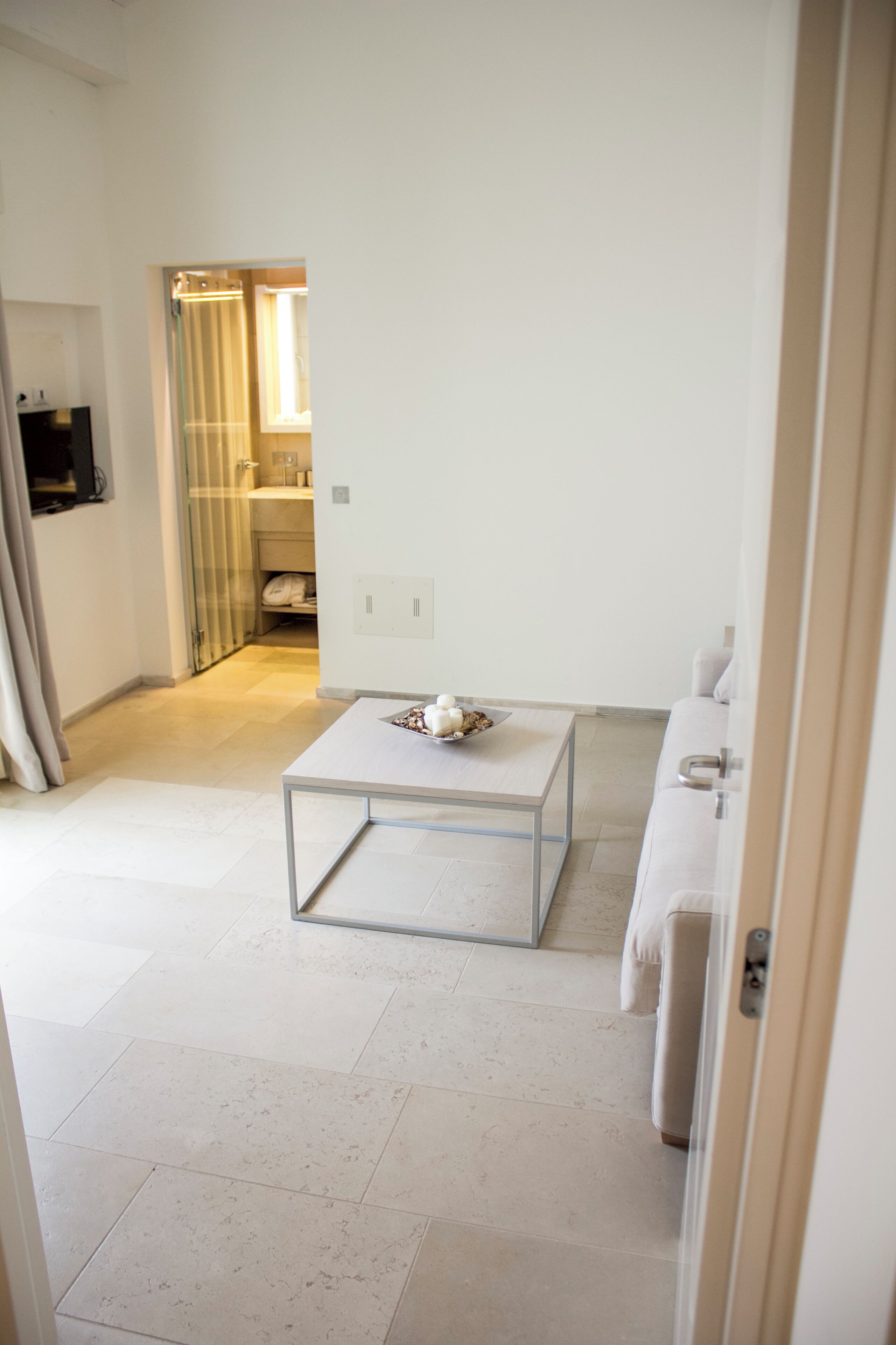 A modern, minimalist living room with a glass coffee table, a white sofa, and a visible bathroom in the background.
