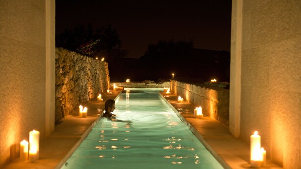 A person in a narrow, illuminated water channel with candles on either side, suggesting a spa or relaxation area.