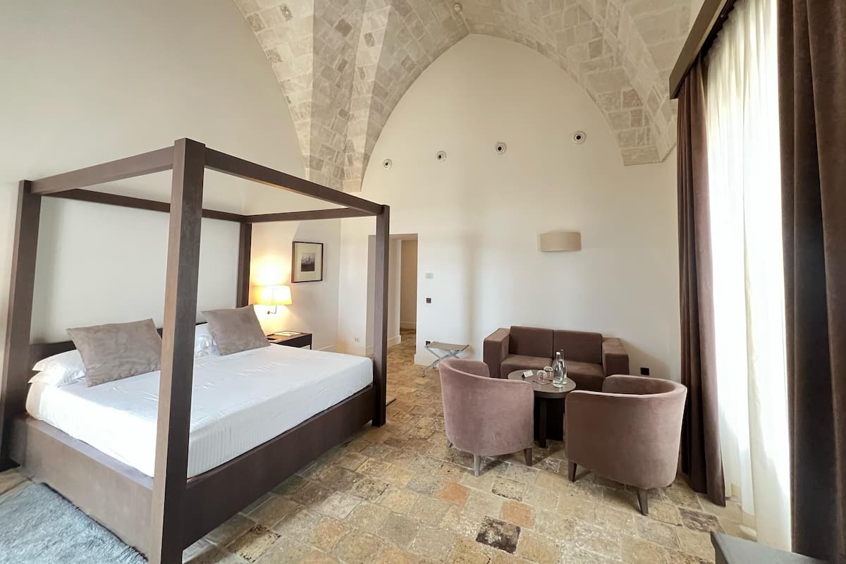 Una cama con dosel, una mesita de noche con lámpara, un sofá y una pequeña mesa con sillas en una habitación con suelo de piedra y un techo alto en arco.